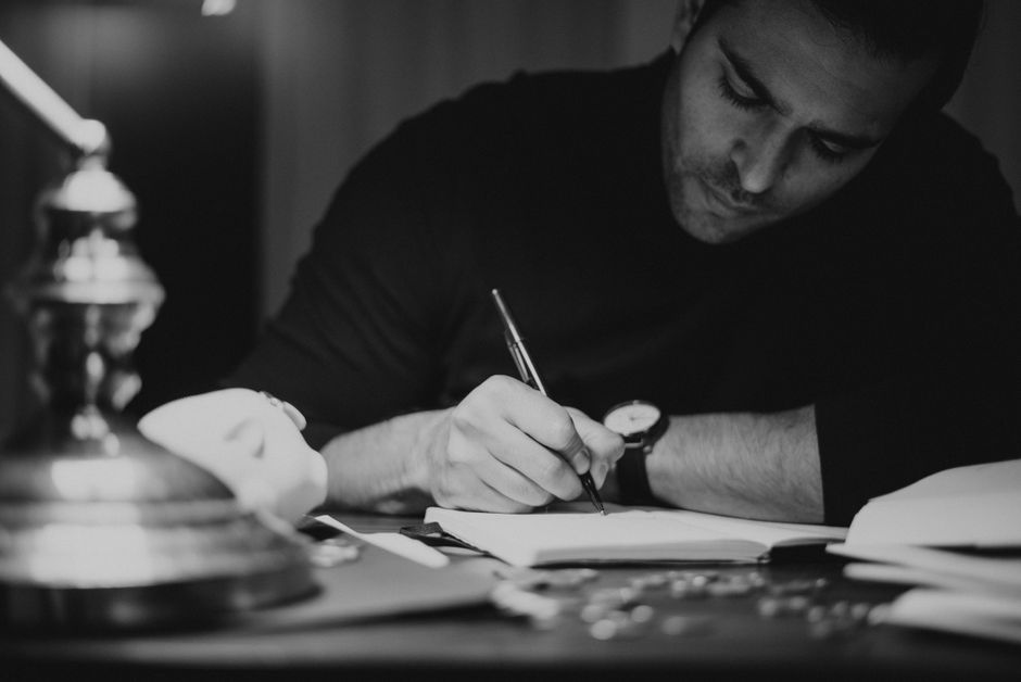 Crop concentrated male write in dark clothes taking notes in notepad while sitting at desk in home office