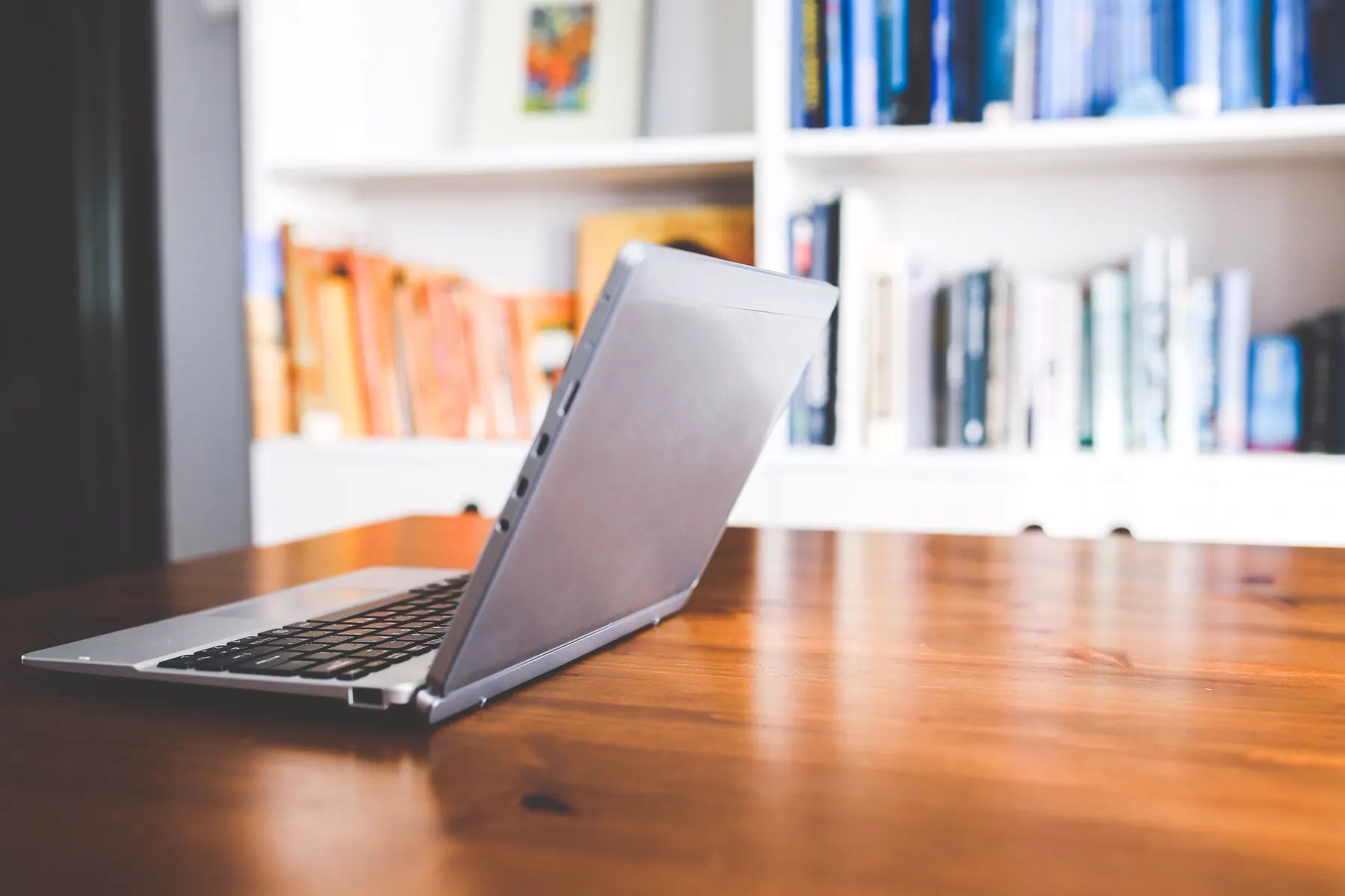 Laptop, stacked books and an open notebook on a wooden desk in front of bookshelves